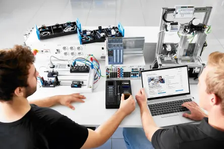 Two learners work with automation training systems while reviewing data on a laptop.