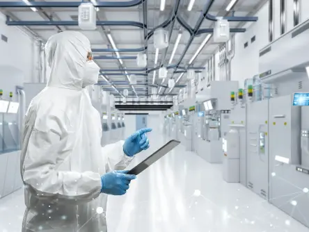 Technician in cleanroom suit tests semiconductor production plant. At top, FOUPs on a transport system.