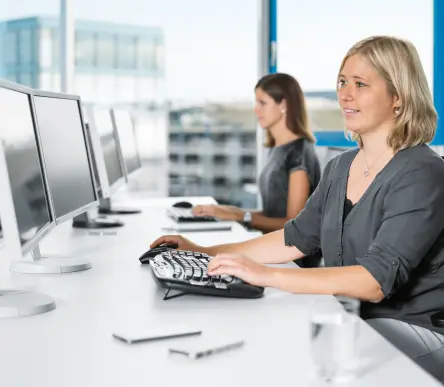 两位女士在配有落地窗的明亮办公室里，对着台式电脑办公