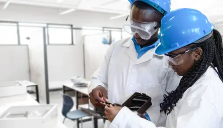 Worker wearing a white hard hat, protective gloves and a blue work coat installs or adjusts electrical fixtures on a wall; the worker is leaning forward and focused on the task, with wiring and mounting elements visible.