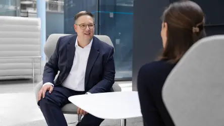 Two people in conversation in a modern office area, with one person in a suit smiling while seated across from a colleague.