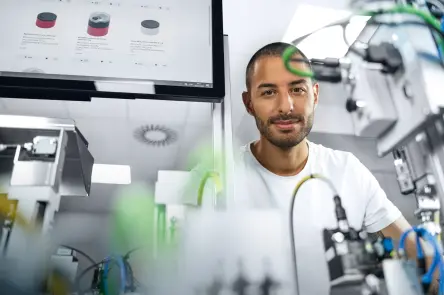 A man in a white T-shirt observes an automated production setup in a room. In the foreground are blurred pneumatic and electrical components, tubing, and sensors mounted on metal fixtures; a touchscreen monitor displaying product images is visible to the left.